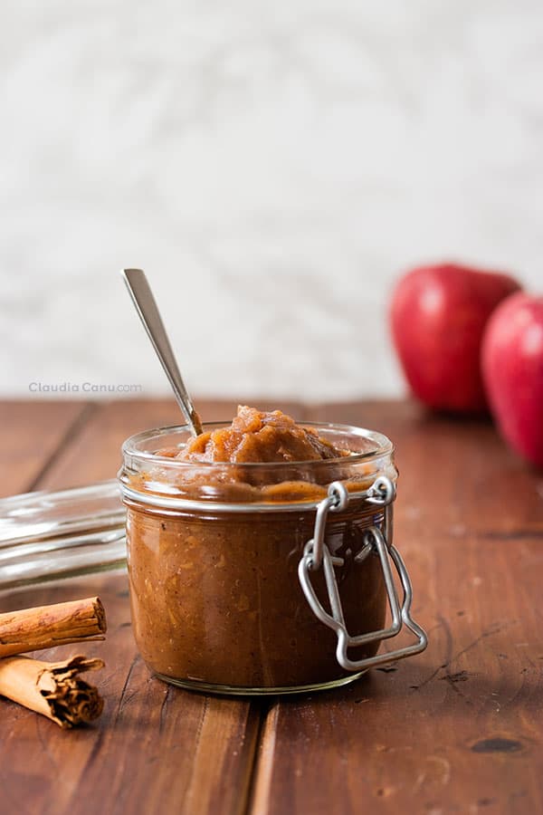 Apple sauce in a jar with spoon sticking out set on a wood surface with cinnamon sticks and apples scattered around.