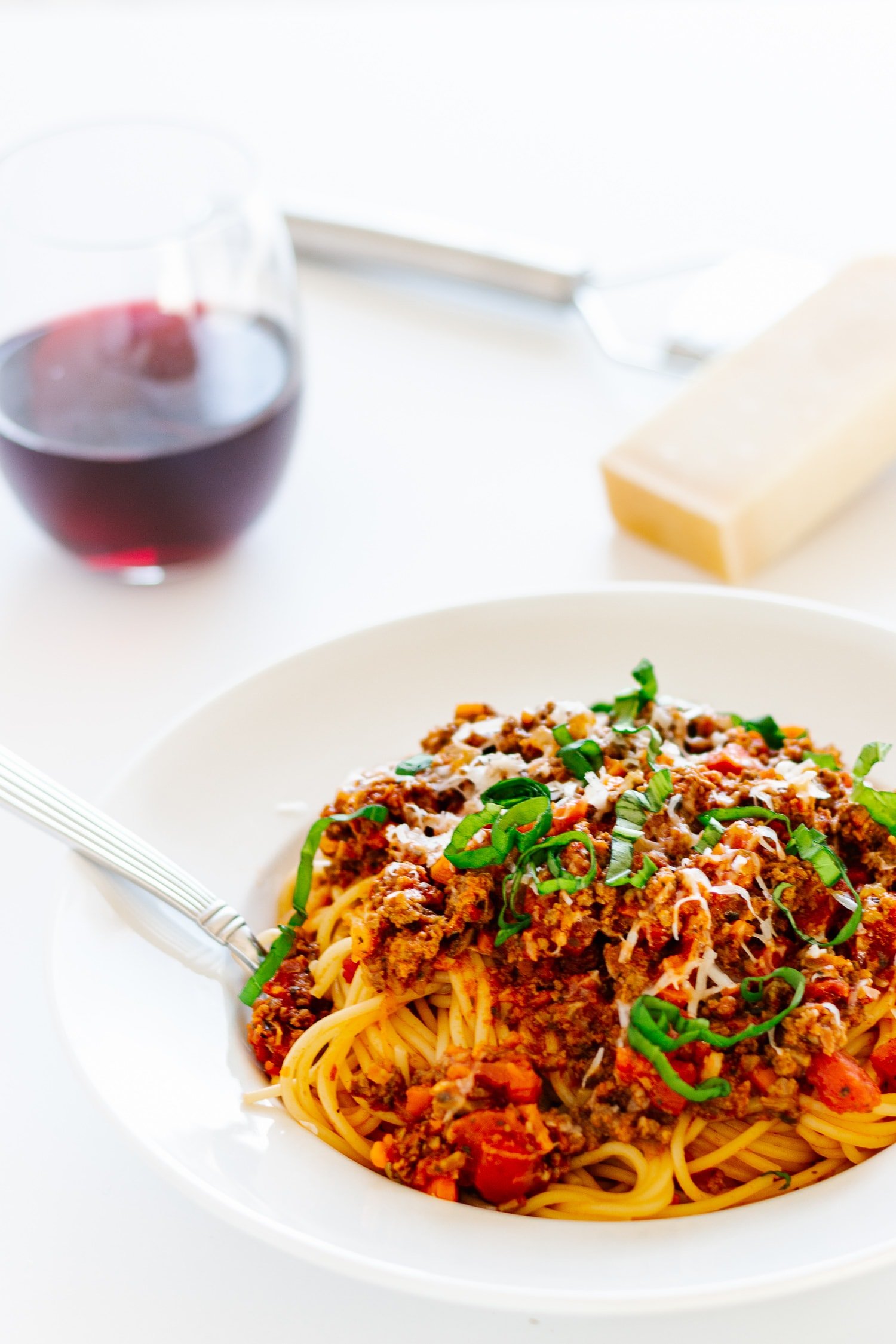 White bowl of spaghetti bolognese with basil scattered on top and a glass of red wine and chunk of parmesan and a grater in the background