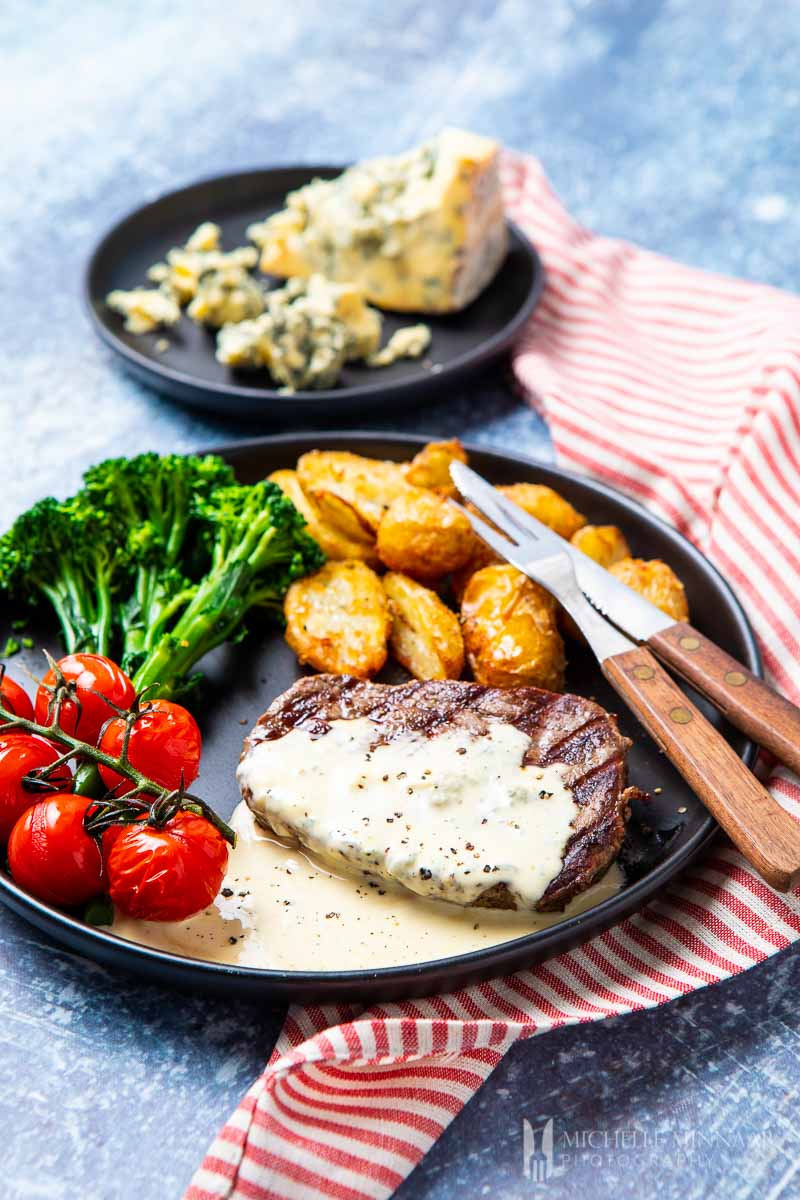 Grey plate containing steak covered in blue cheese sauce, roasted tomatoes on stem, steamed broccoli and roasted potatoes with fork and knife resting on the edge.  Background contains a second plate with a chunk of blue cheese.