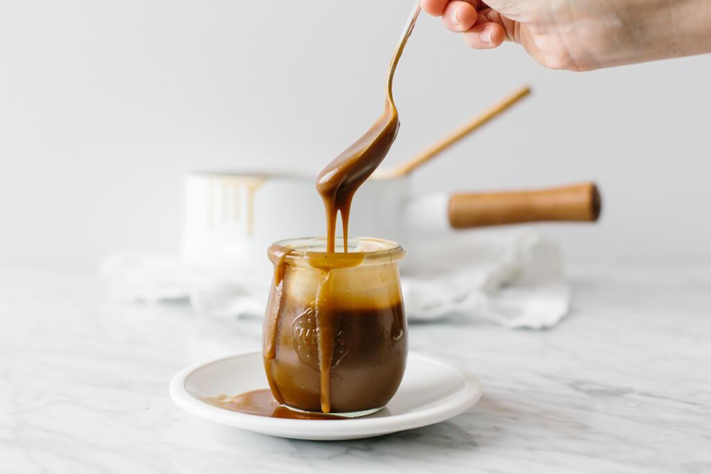 Jar of salted caramel sauce set on a white plate.  Spoon lifting caramel sauce out and cooking pot in the background.