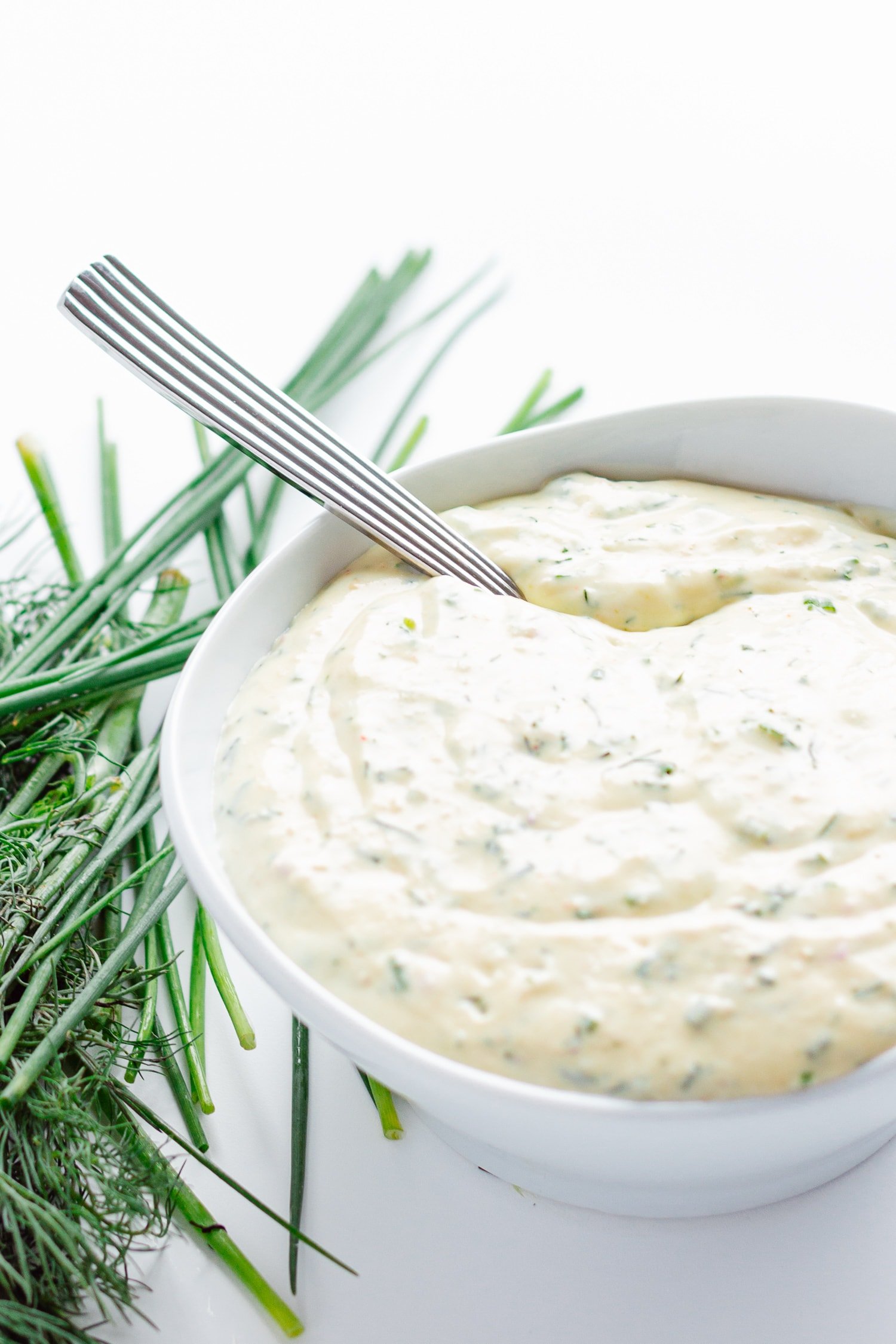 White bowl of creamy herb sauce/dip with spoon handle sticking out.  Scattering of dill and chives on the side.