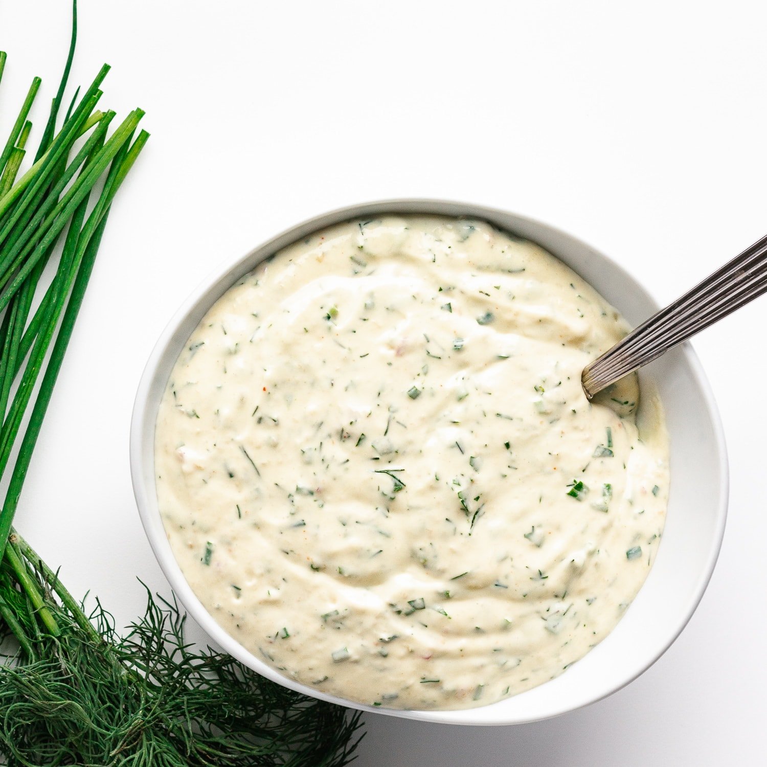 Overhead photo of creamy herb sauce/dip in a white bowl with silver spoon handle sticking out.  Scattering of chives and dill to the left of the bowl.