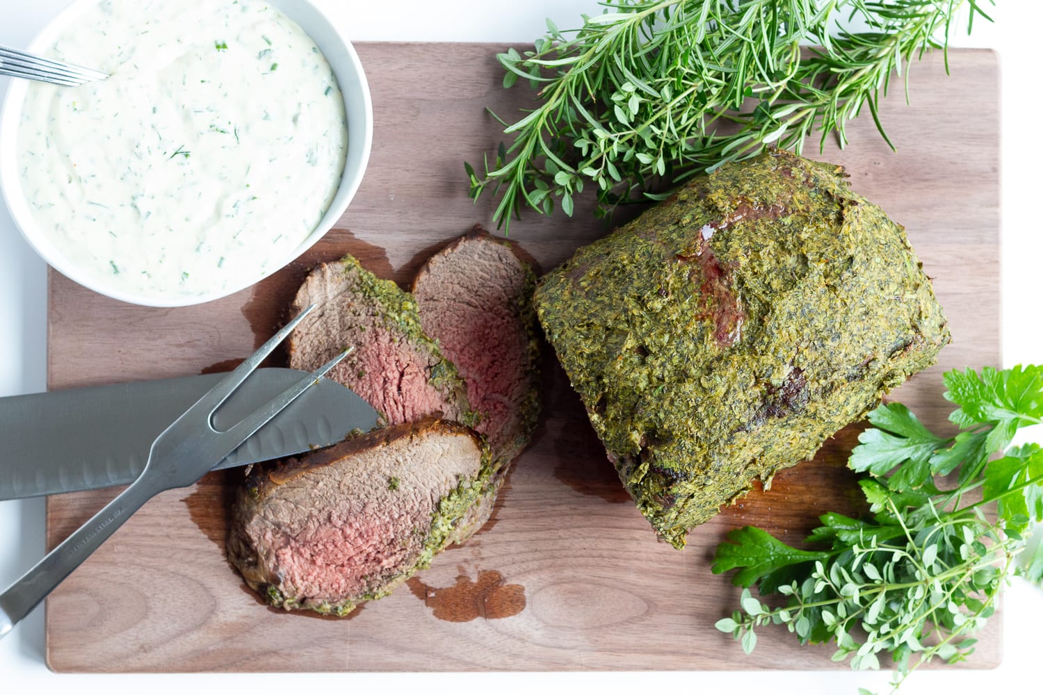 Overhead photo of herb mustard crusted roast beef on a wooden carving board with 3 slices carved off.  There are also fresh herbs, bowl of creamy herb sauce and carving utensils laid out on the board.