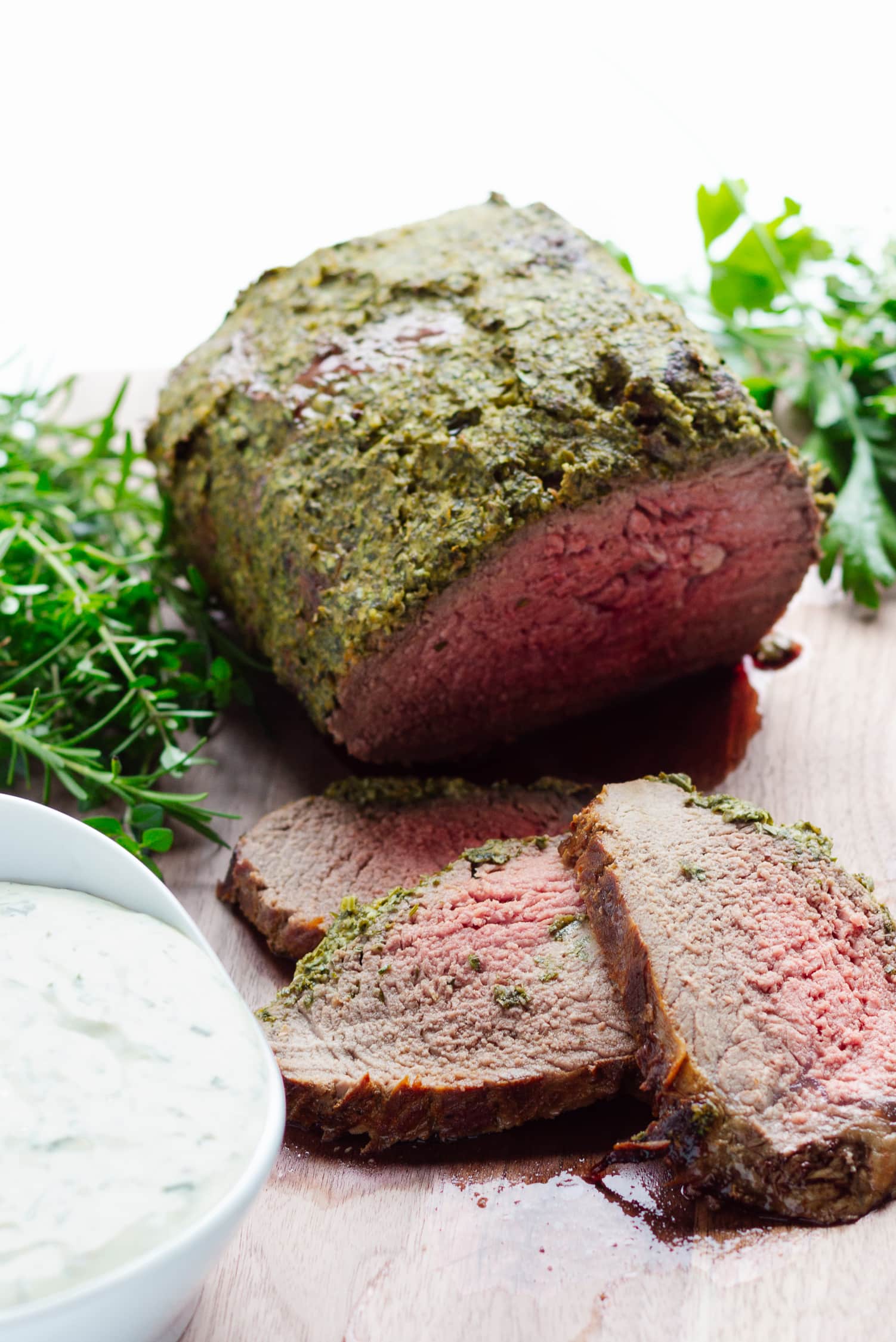 Herb Crusted Roast Beef on a wooden cutting board with 3 slices carved off in front.  Also a bowl of creamy herb sauce and fresh herbs on the board.