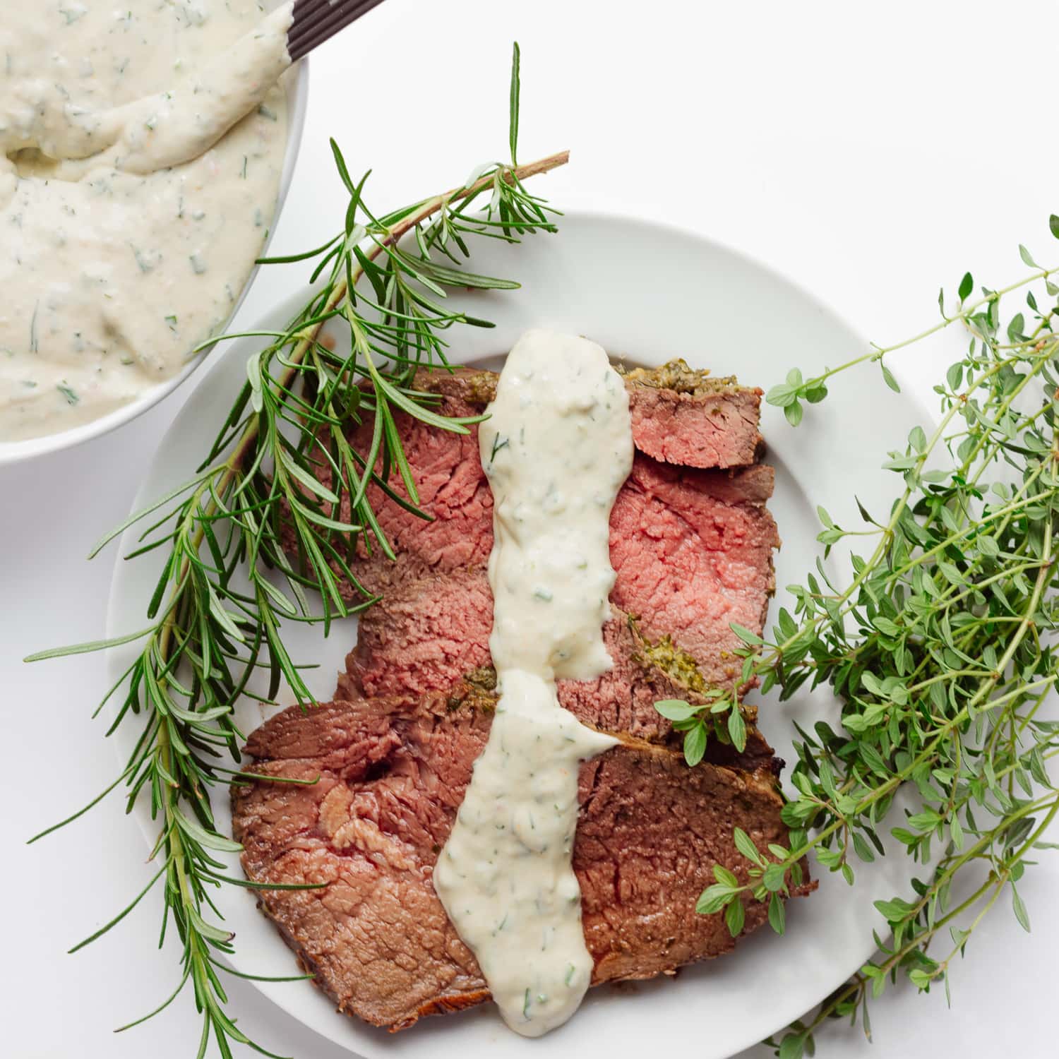 Slices of roast beef on a white plate with a drizzle of creamy herb sauce down the middle.  There is also a bowl of the same sauce in the corner and a sprig of rosemary and thyme on each side of the plate.