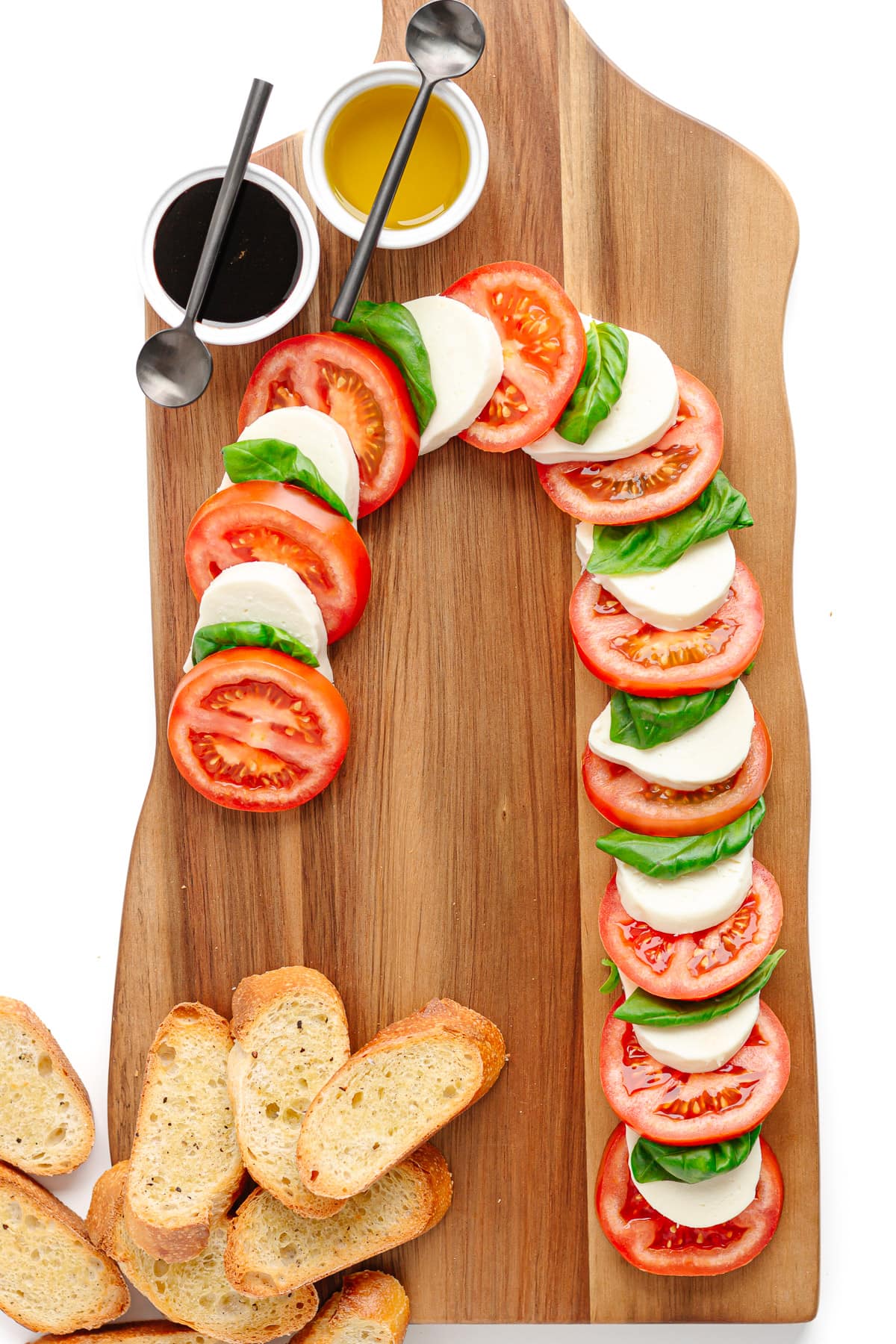 Tomato slices, fresh mozzarella slices and basil leaves arranged into a candy cane shape on a wooden board along with crostini and two small bowls containing balsamic glaze and olive oil.