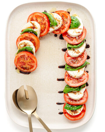 A caprese salad arranged in the shape of a candy cane on a cream colored rectangular serving platter with gold salad servers.