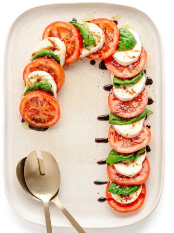 A caprese salad arranged in the shape of a candy cane on a cream colored rectangular serving platter with gold salad servers.