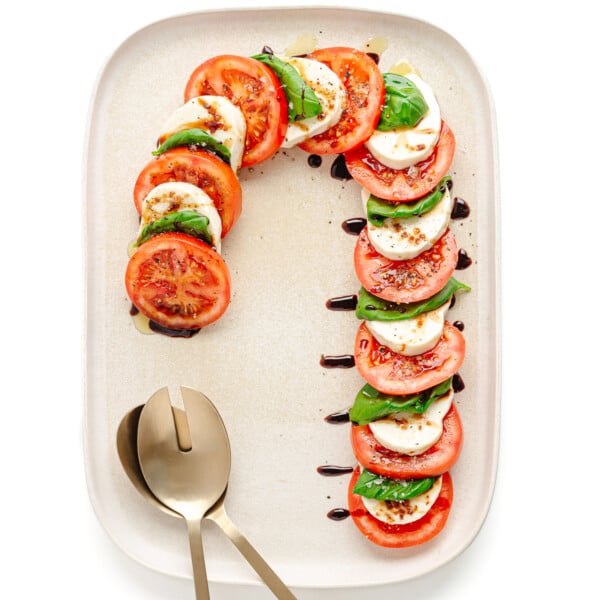 A caprese salad arranged in the shape of a candy cane on a cream colored rectangular serving platter with gold salad servers.