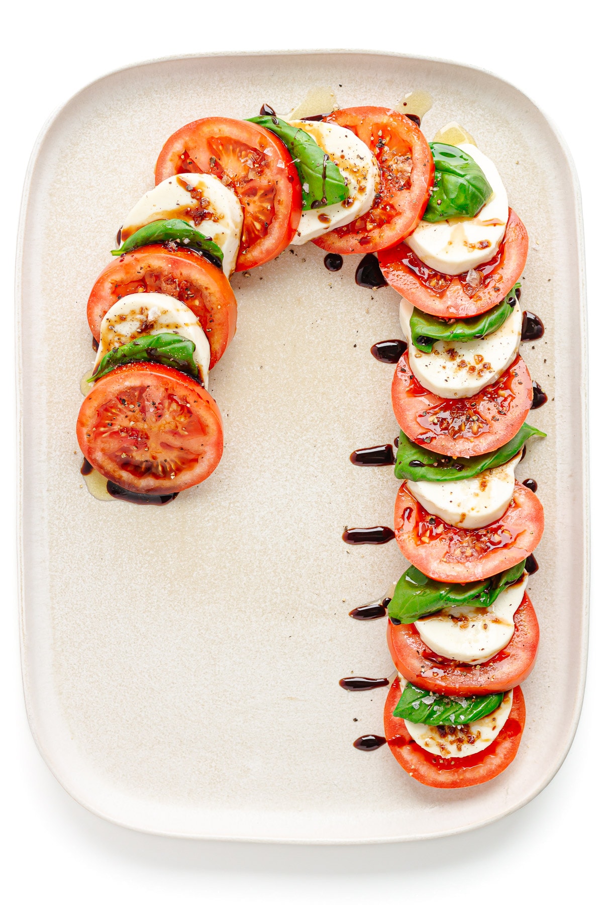 A Christmas caprese salad arranged in the shape of a candy cane on a cream colored rectangular serving platter.