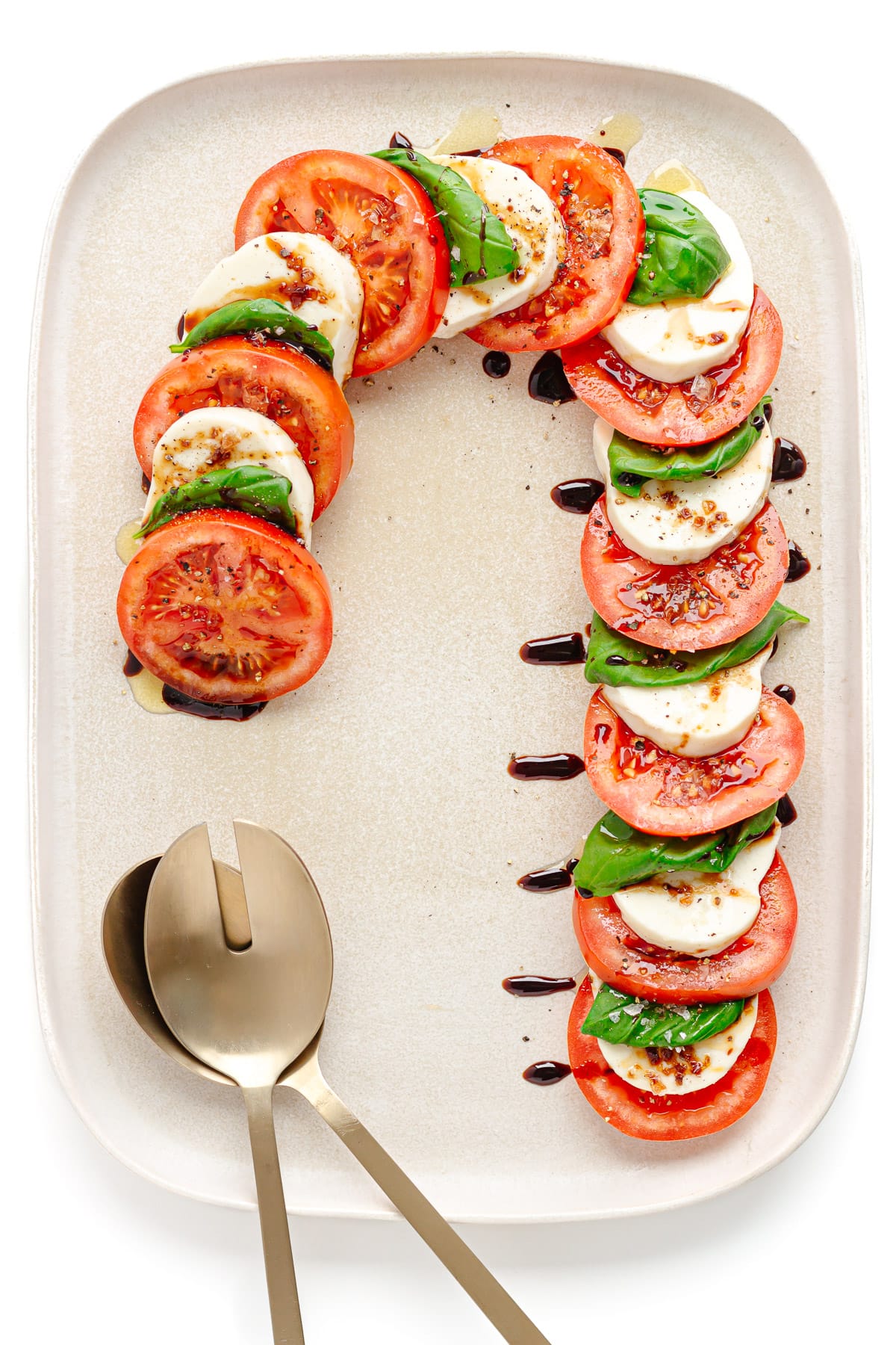 Caprese salad arranged in the shape of a candy cane on a cream colored platter with salad servers.