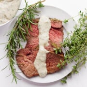 A few slices of herb mustard crusted roast beef on a white plate with creamy herb sauce drizzled over top.