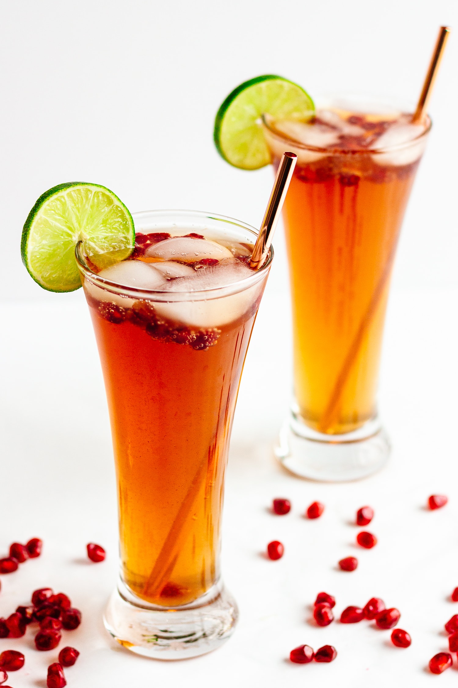 Two glasses of pomegranate lime spritzer on a white background with pomegranate arils sprinkled around