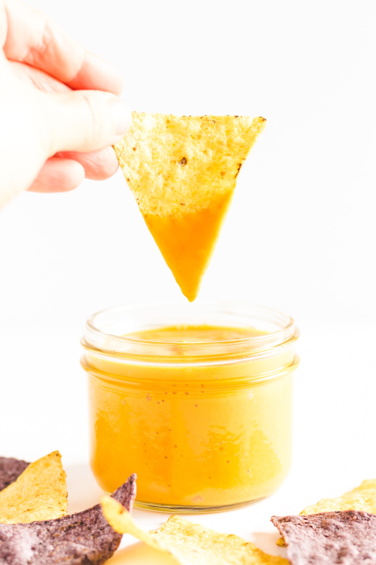 Hand holding tortilla chip being dipped into a jar of homemade vegan and nut free nacho cheese sauce.