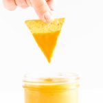 Tortilla chip being dipped into a jar of vegan + nut free nacho cheese sauce