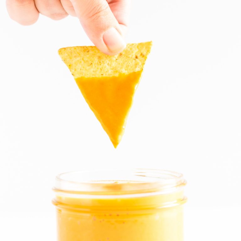 Tortilla chip being dipped into a jar of vegan + nut free nacho cheese sauce