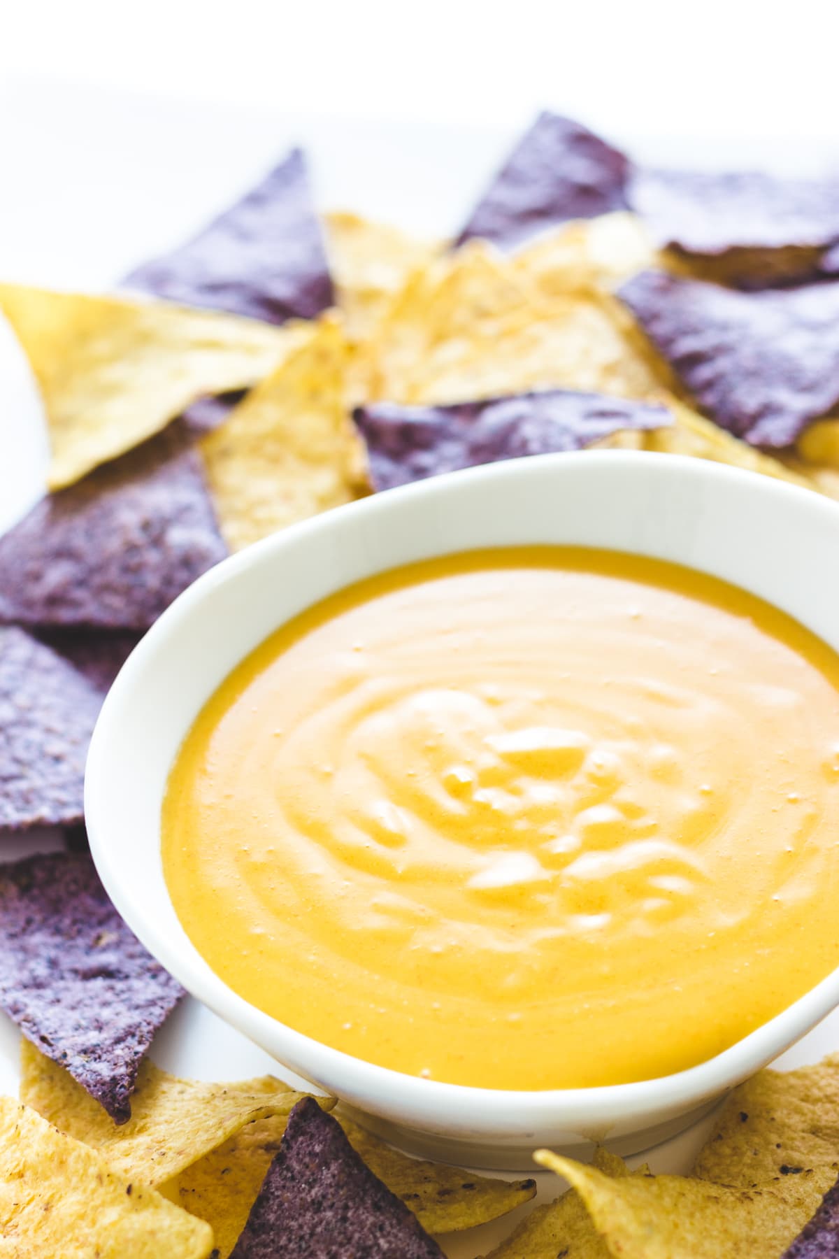 Homemade nacho cheese sauce in a white bowl surrounded by yellow and blue corn tortilla chips