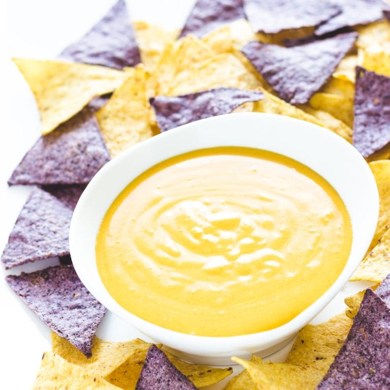 Homemade nacho cheese sauce in a white bowl surrounded by blue and yellow tortilla chips.