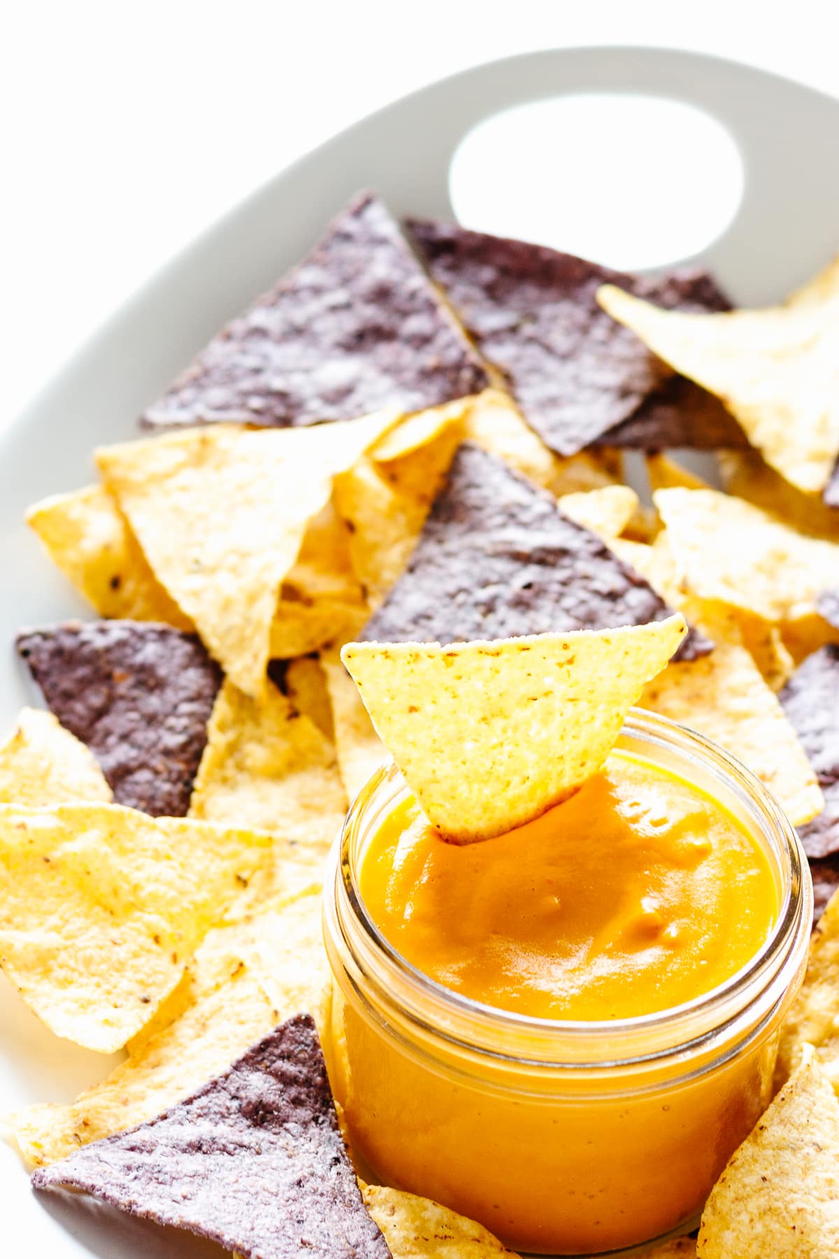 White platter of yellow and blue corn tortilla chips and a jar of homemade vegan nacho cheese.