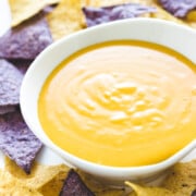 White bowl of homemade nacho cheese sauce surrounded by blue and yellow corn tortilla chips.