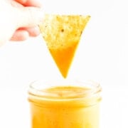 Hand dipping tortilla chip in a glass jar of veggie nacho cheese sauce.