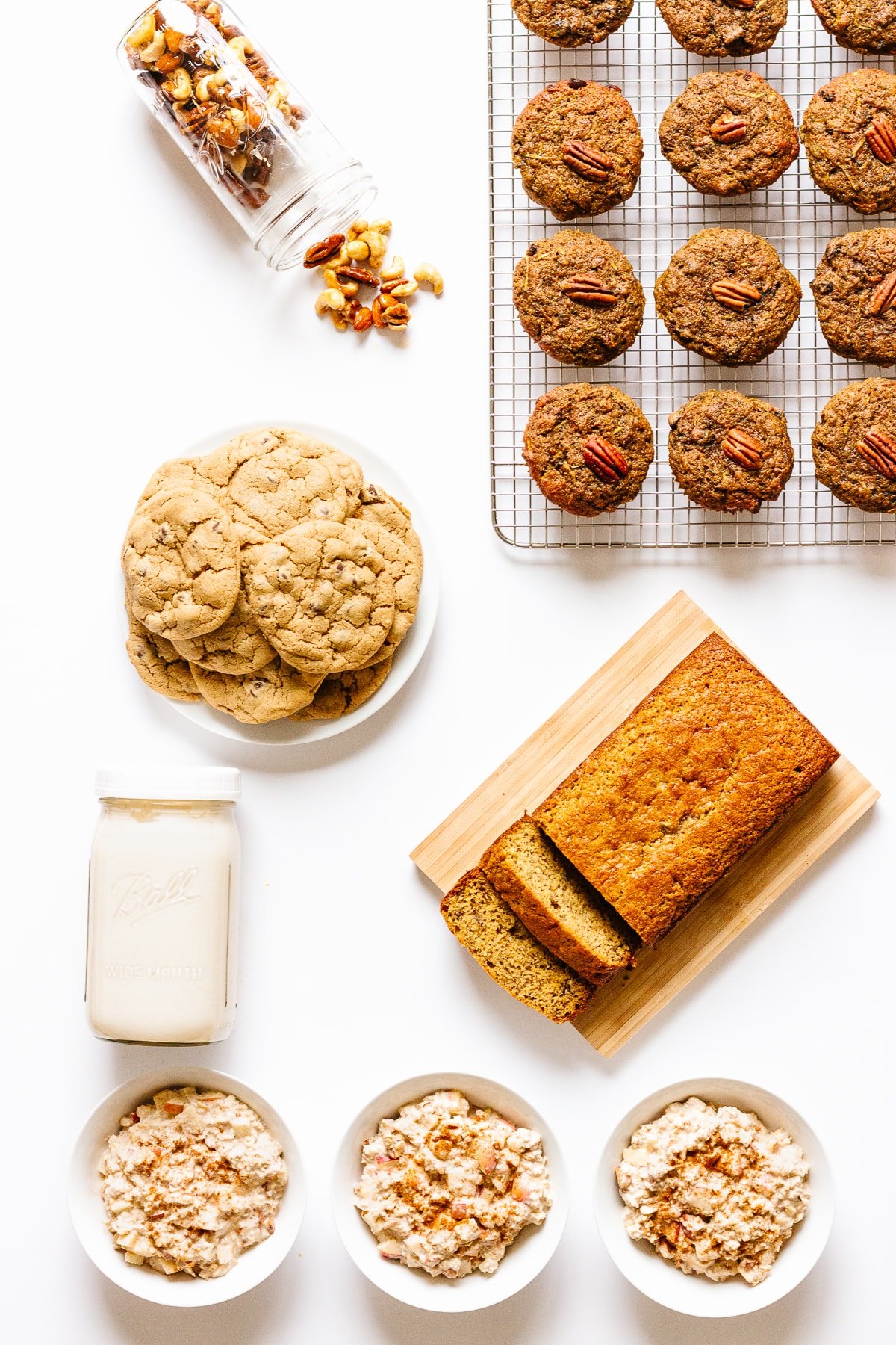 Overhead photo of muffins, spiced nuts, chocolate chip cookies, banana bread, jar of cashew cream and 3 bowls of Bircher Muesli all set on a white background.
