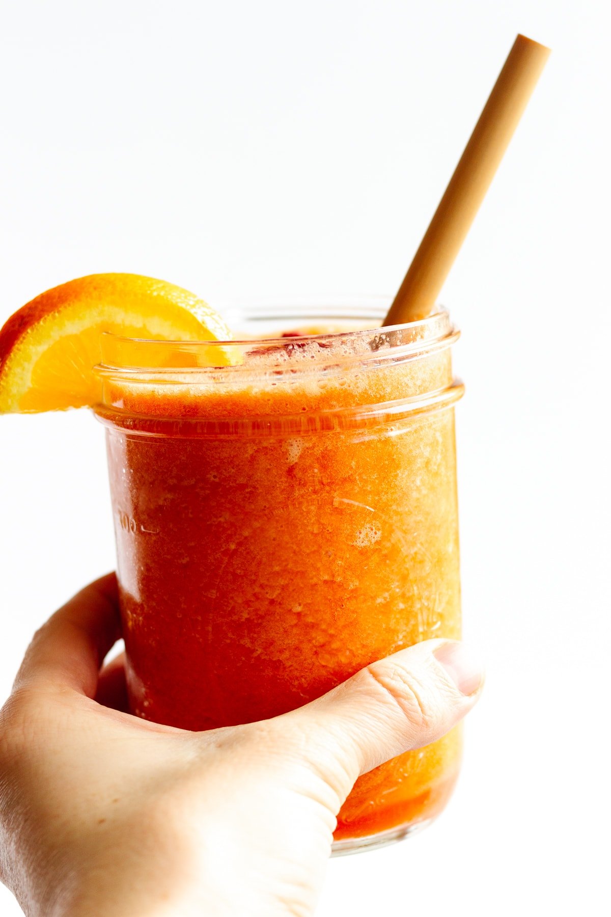 Hand holding mason jar filled with Vitamin C Smoothie with an orange slice on the edge and bamboo straw.