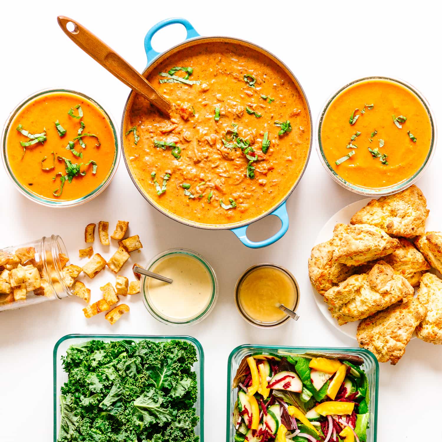 Overhead photo of a pot of garden bolognese, 2 containers of tomato soup, croutons pouring out of a jar, 2 jars of salad dressing, plate of biscuits and 2 containers of salad all set on a white background.