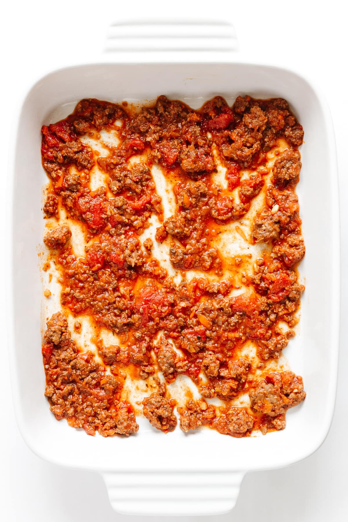 Thin layer of Bolognese Sauce spread on the bottom of a white casserole dish.