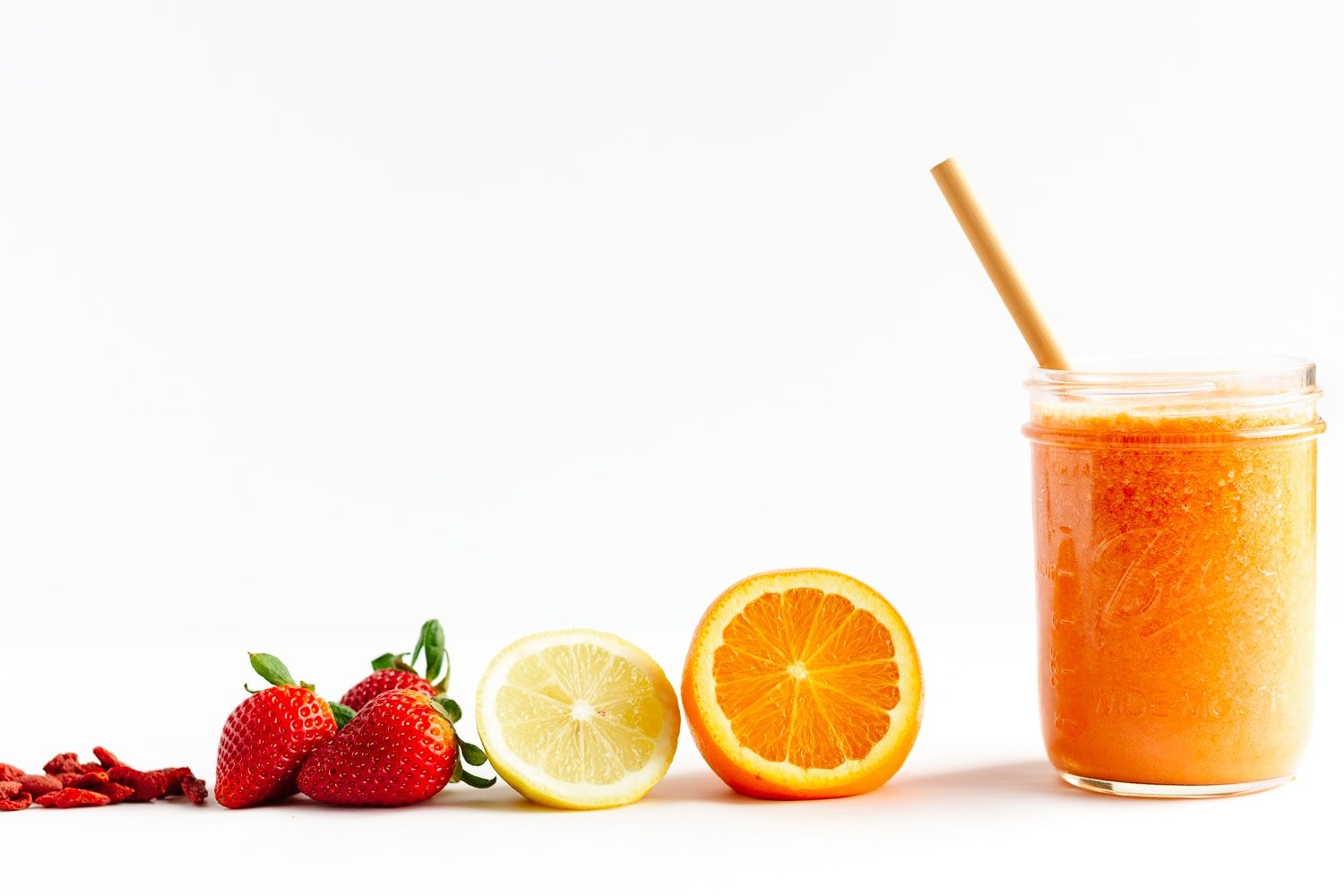 Goji berries, strawberries, lemon, orange and glass of Vitamin C Smoothie lined up on a white background.