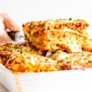 Slice of lasagna being lifted from a whole lasagna in a white casserole dish.