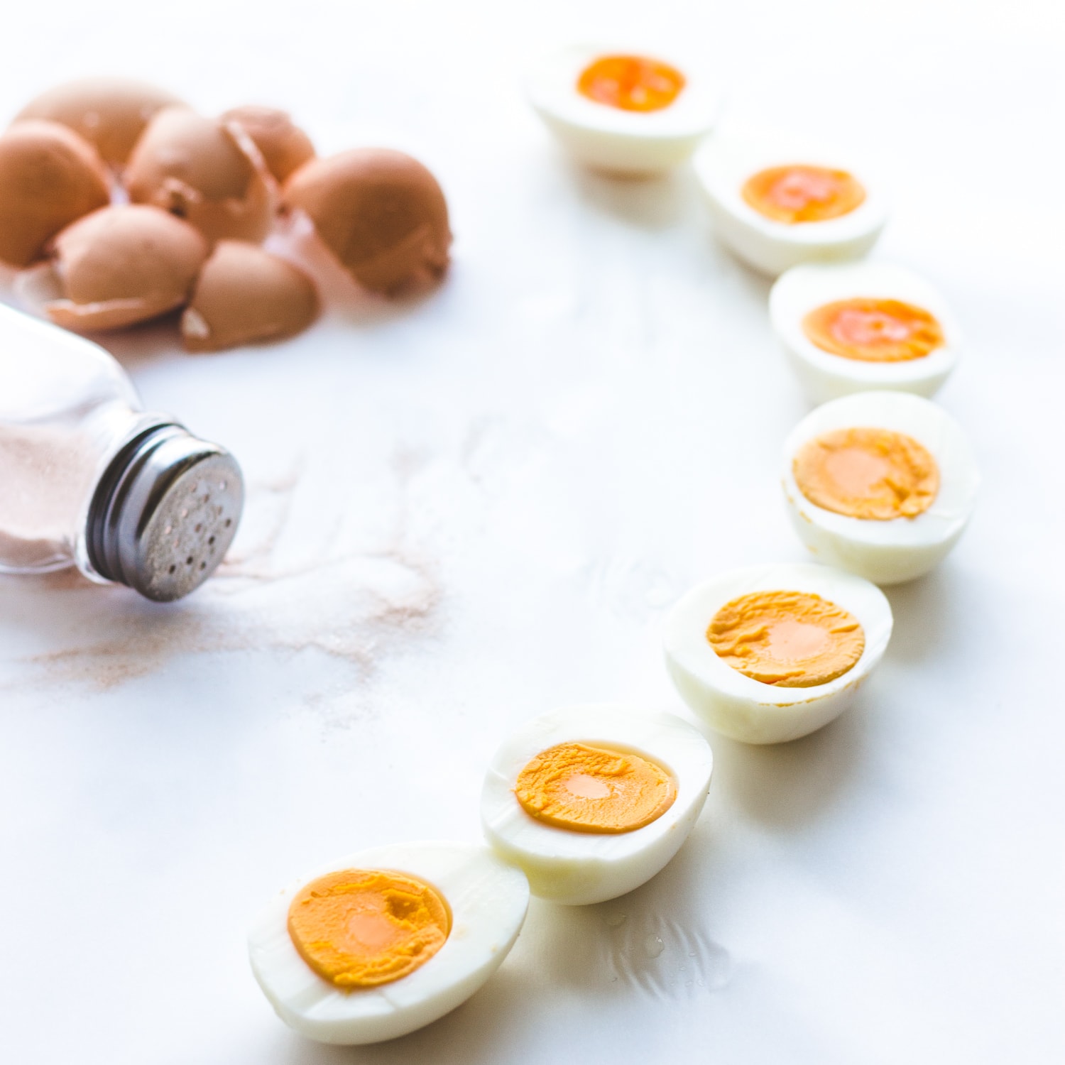 Hard boiled eggs lined up in a curved shape with egg shell and salt shaker in the corner.