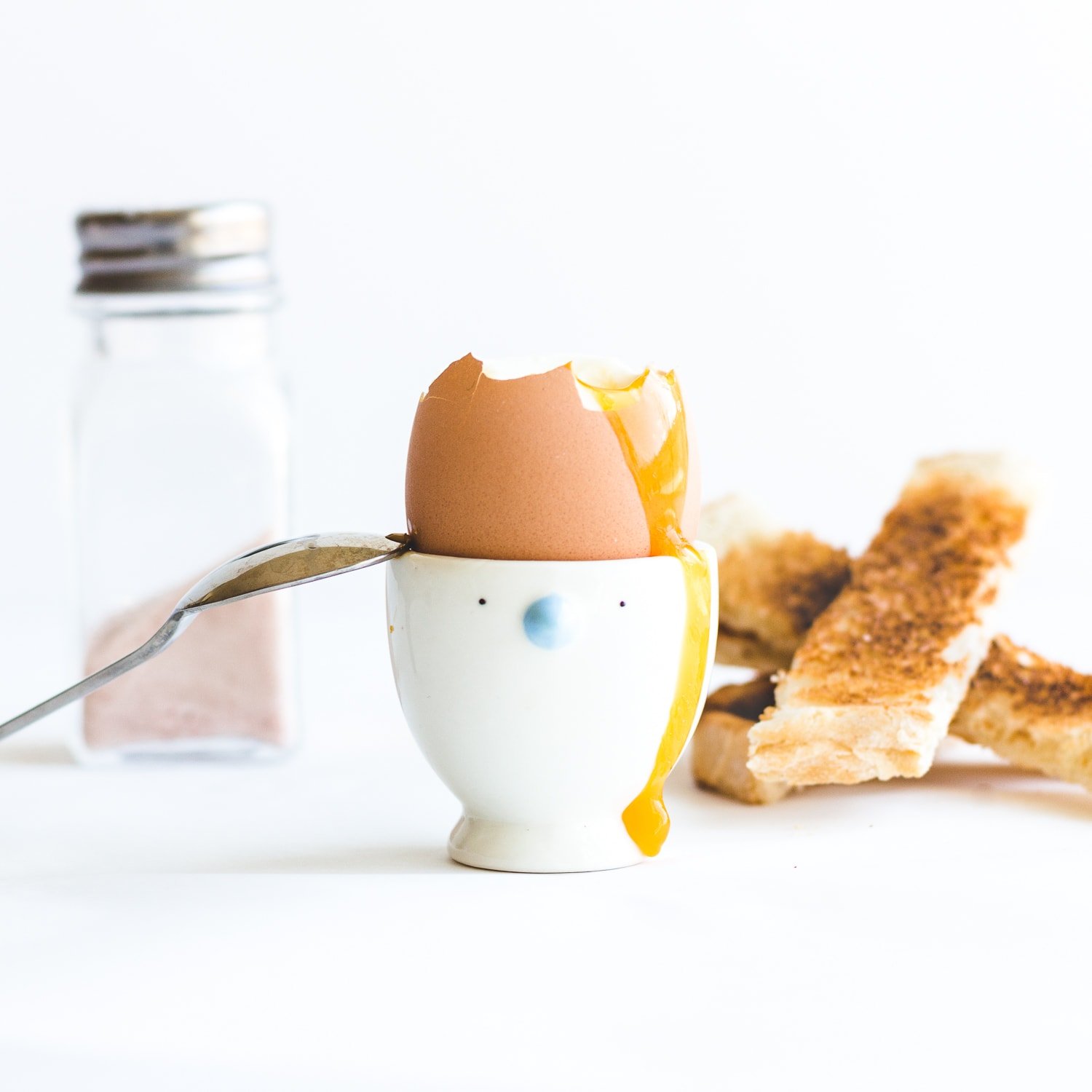 Soft boiled egg in egg cup with top off, yolk running down the side and spoon leaning on the edge.  Pile of toast soldiers and salt shaker in the background.