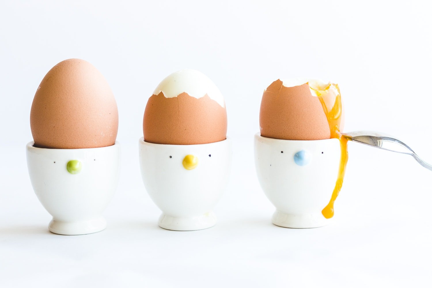 Three soft boiled eggs in egg cups with yolk running down the side of the last one and a spoon resting against it