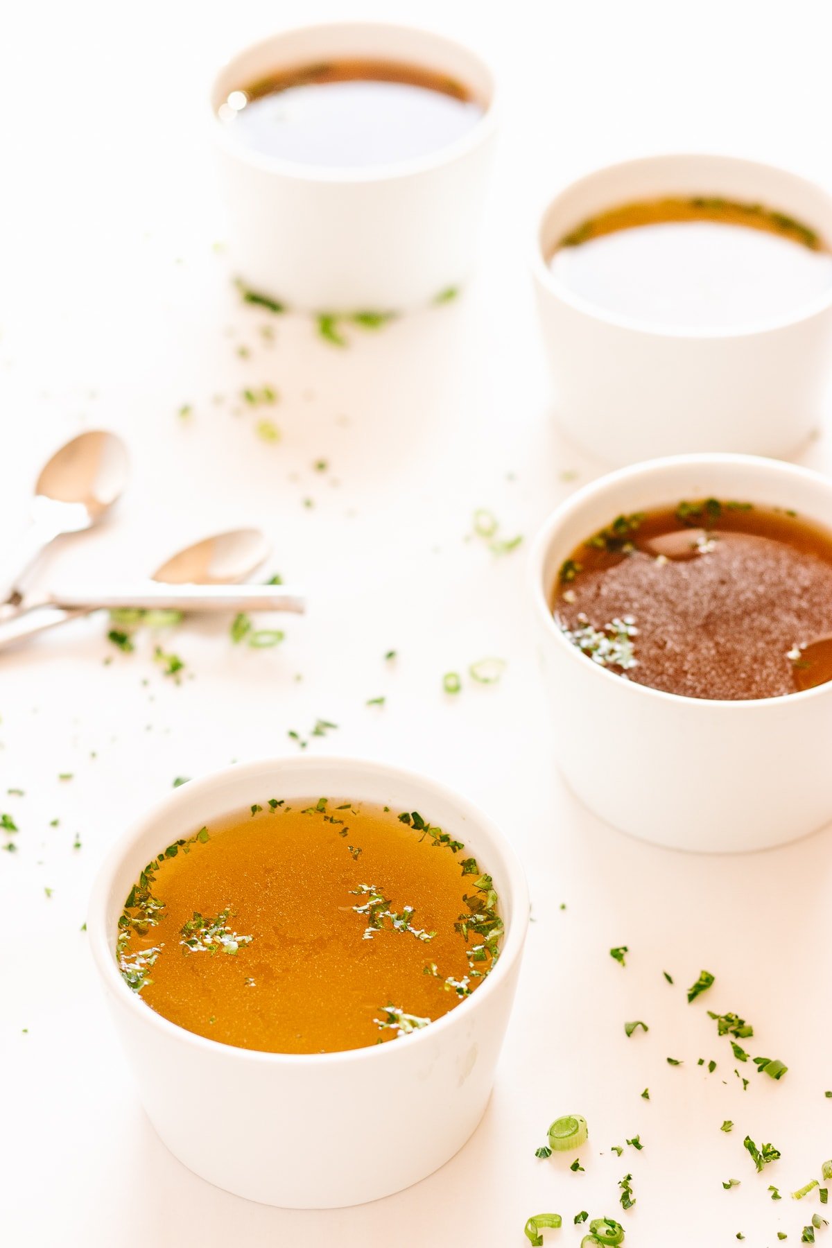 Four white bowls of broth on a white background with scattered herbs and spoons beside them