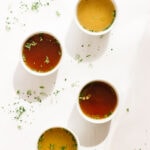 Four white bowls of broth on a white background with scattered chopped herbs.