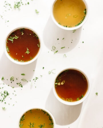 Four white bowls of broth on a white background with scattered chopped herbs.