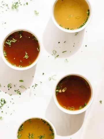 Four white bowls of broth on a white background with scattered chopped herbs.