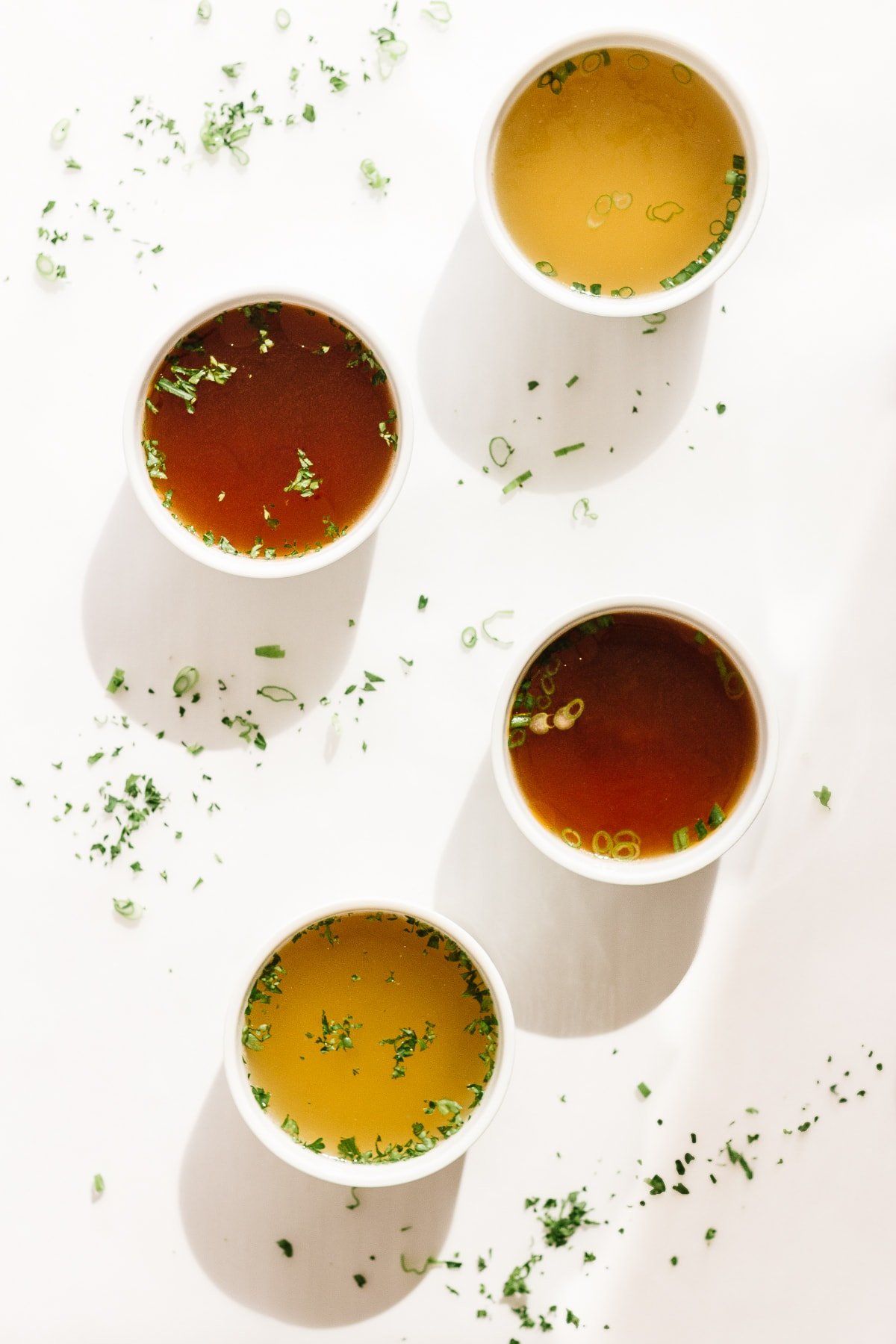 Four white bowls of broth set on a white background with chopped herbs scattered.