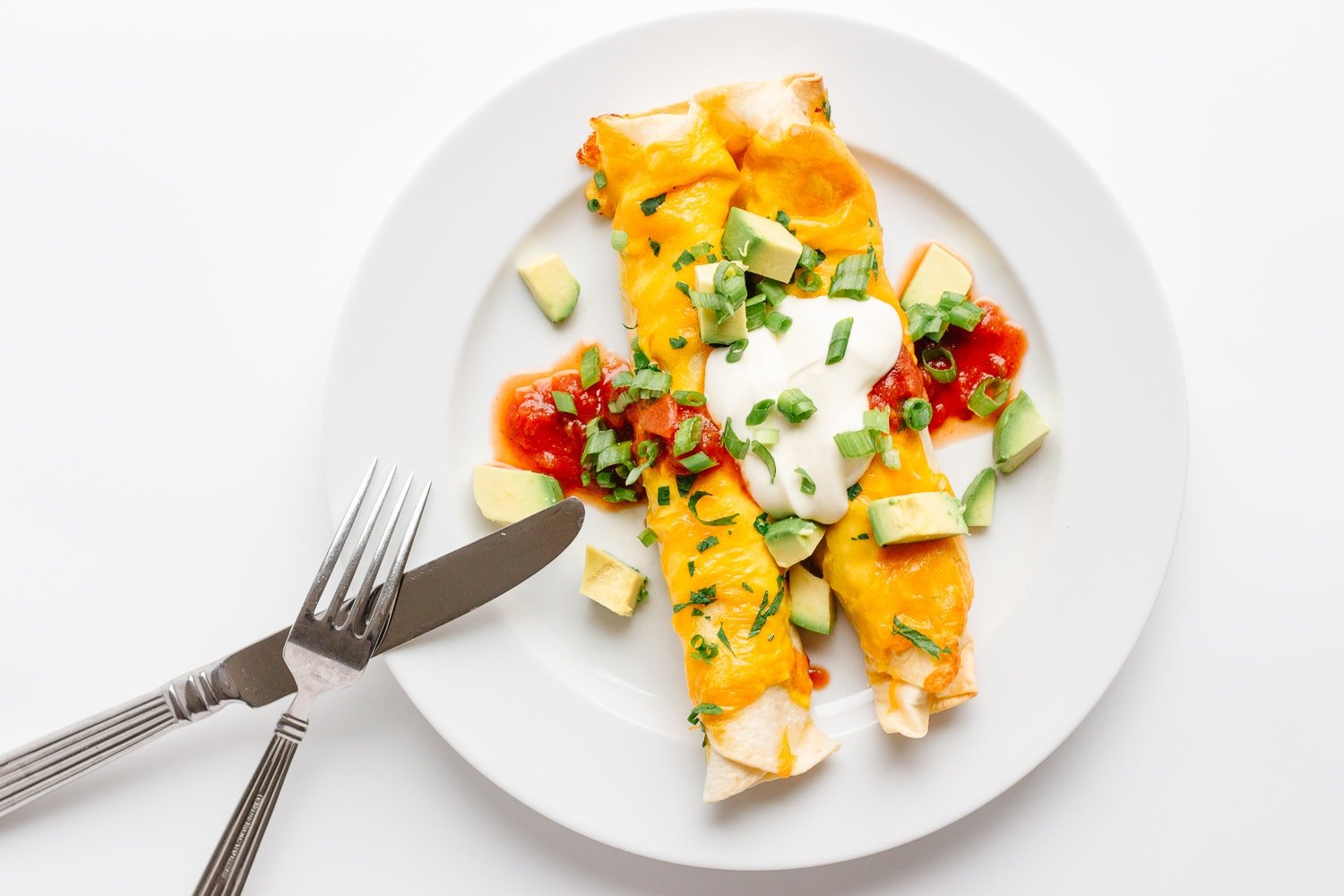 Two chicken enchiladas on a white plate with fork and knife leaning on the edge.