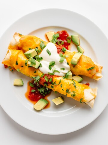 Two cheesy chicken enchiladas on a white plate and garnished with salsa, sour cream, cubed avocado and chopped green onion.