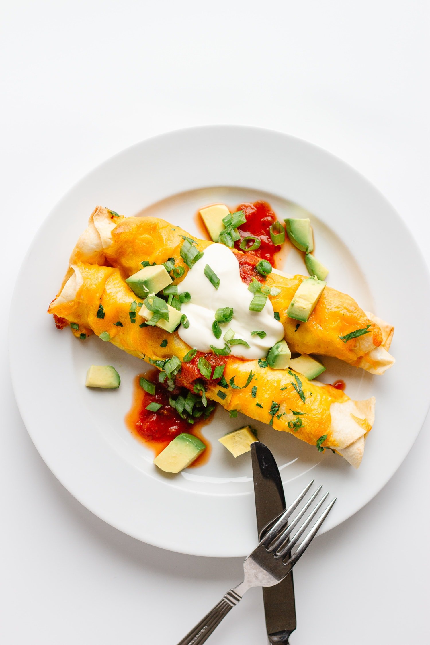 Two cheesy chicken enchiladas on a white plate garnished with cubed avocado, salsa, sour cream and chopped green onions.