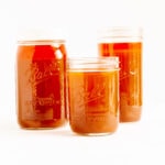 Three different sizes of mason jars filled with beef bone broth (stock) on a white background.