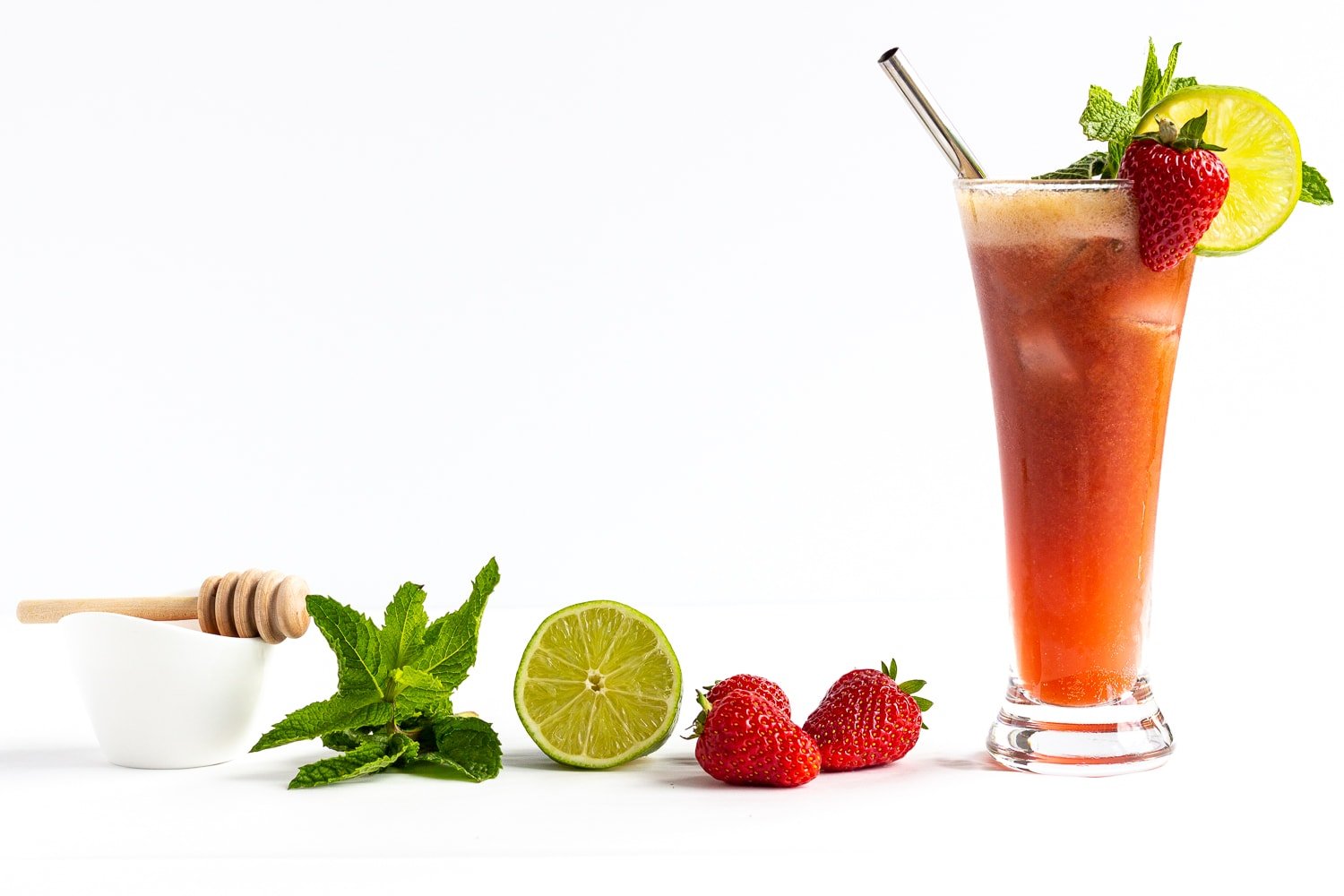 Glass of strawberry mojito mocktail with strawberries, lime, mint and a small bowl of honey next to it.