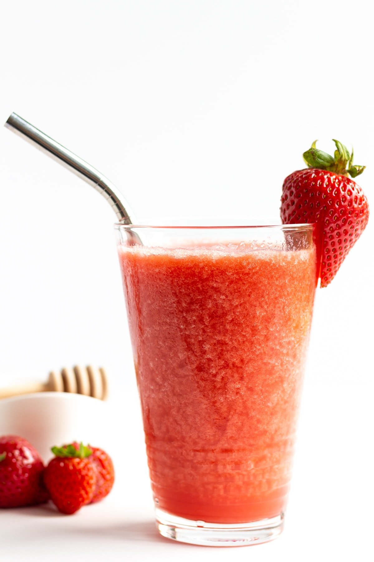 Glass of strawberry slushie garnished with a strawberry and stainless steel straw.