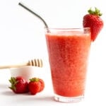 Glass of strawberry slushie with small bowl of honey and a few strawberries in the background.