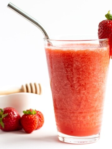Glass of strawberry slushie with small bowl of honey and a few strawberries in the background.
