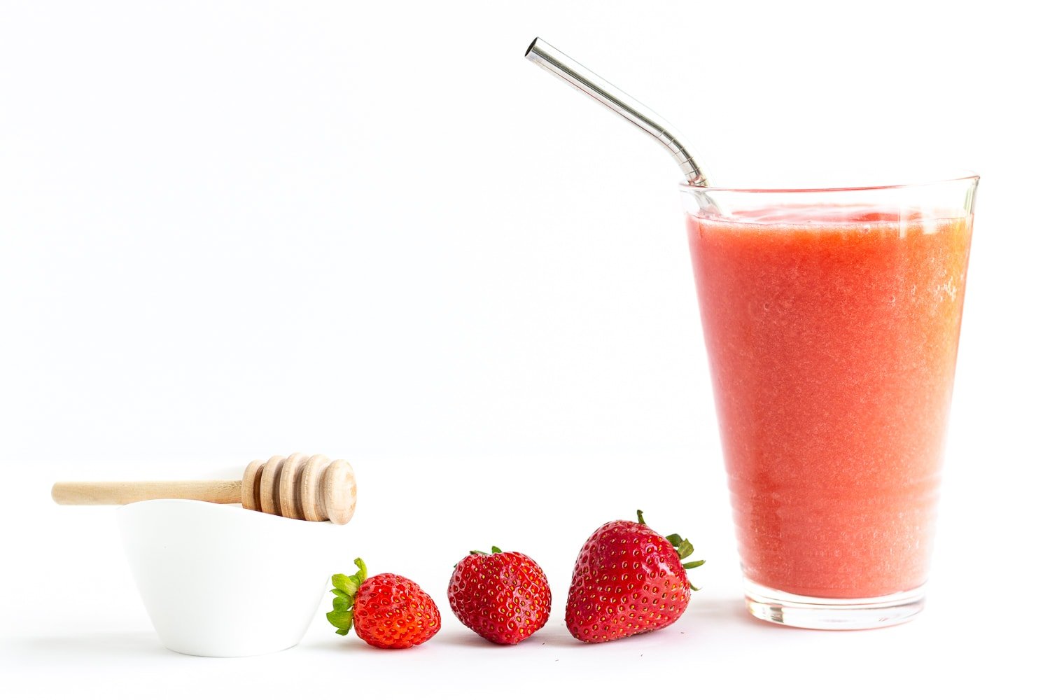 Small white bowl of honey with honey dipper, three strawberries and tall glass of strawberry slushie.