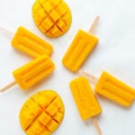 Overhead photo of mango popsicles and fresh mango halves on a white background.