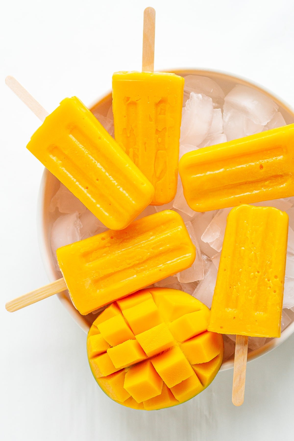 Five mango popsicles and half a fresh mango sitting on a bowl of ice.